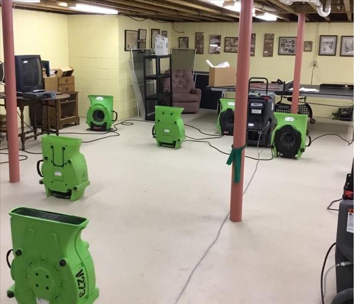 At least six ceiling-facing SERVPRO air movers and two dehumidifiers running in a basement with exposed ceiling joists and pl
