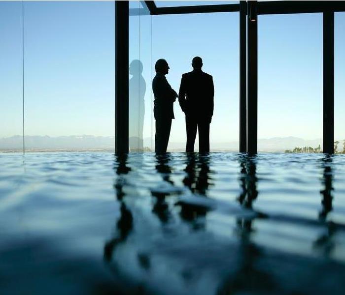 Silhouette of two men standing in floodwater and staring out of large plate glass window of an office